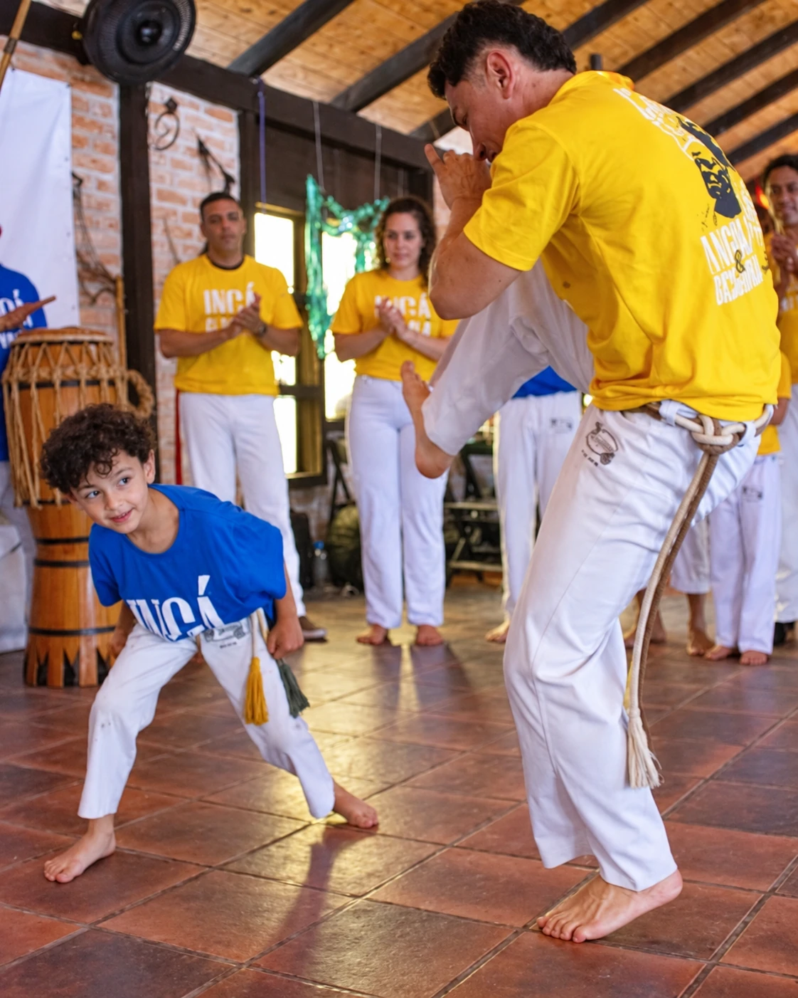 Criança aprende um movimento de capoeira com um adulto na roda do Ingá